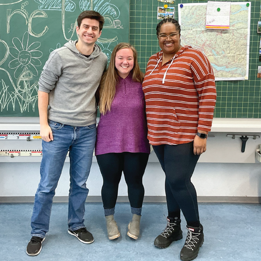 Three students smiling, standing together in front of a chalkboard in a classroom.