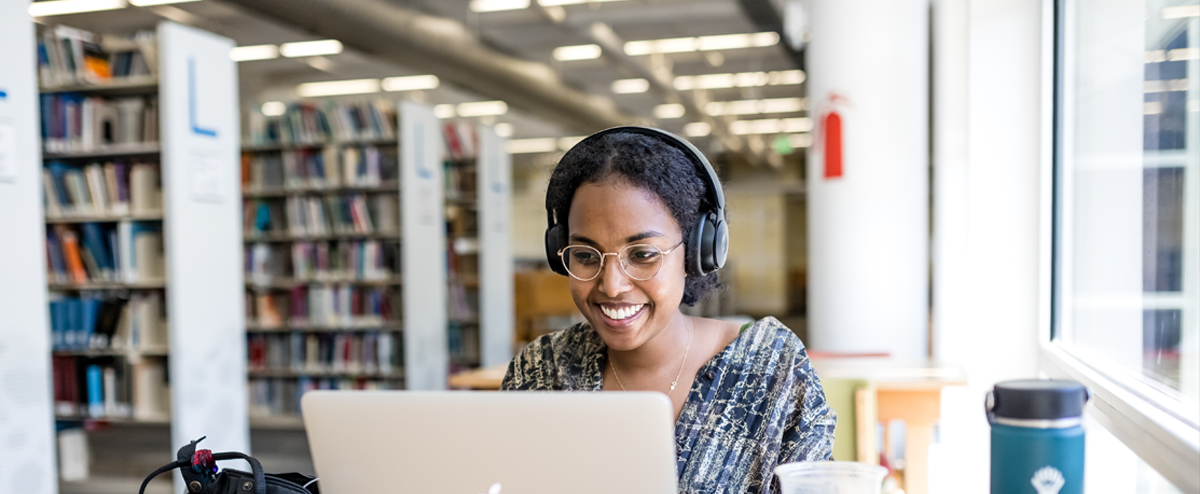 Image of a student sitting in front of a computer with headphones on in the library, smiling.