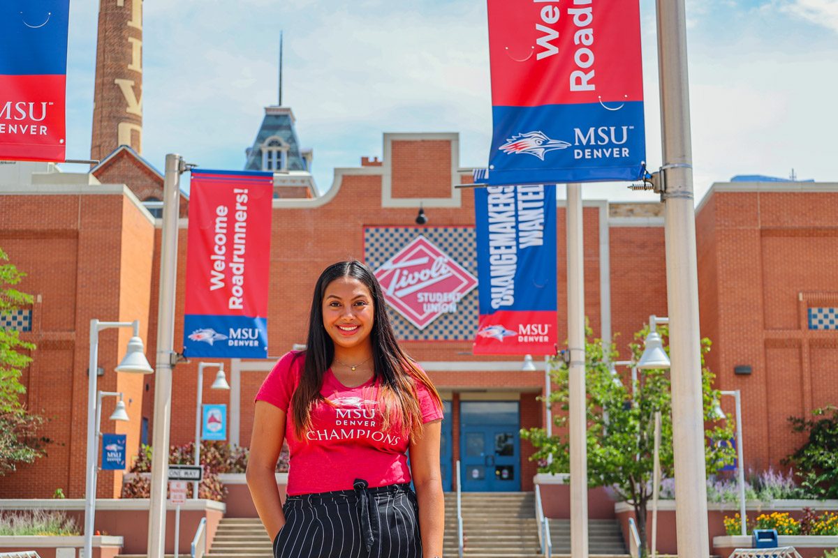 Student Ashley standing in front of the west side of the Tivoli with red and blue MSU Denver signs behind her