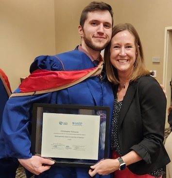 MPAcc graduate (Christopher) in regalia holding diploma frame smiling next to faculty (Jo) at Hooding Celebration