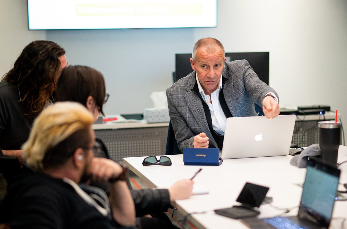 Man in a suit in front of a laptop talking with three students at a table