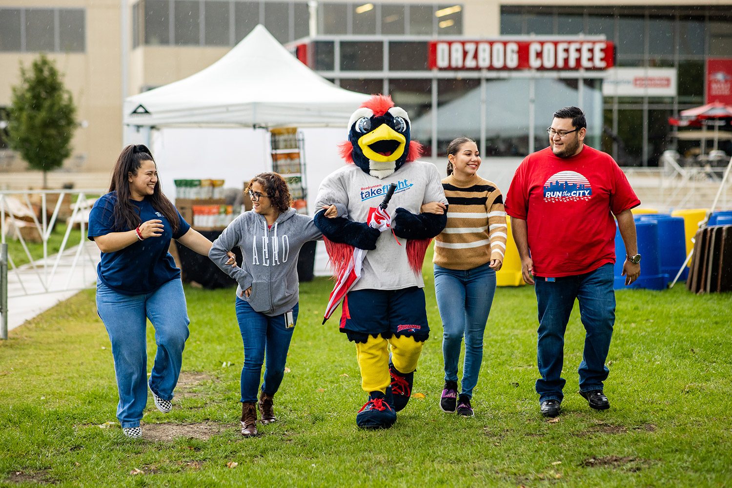 Four alumns walking with Rowdy the Roadrunner at the Homecoming Tailgate on the grass outside of the JSSB building