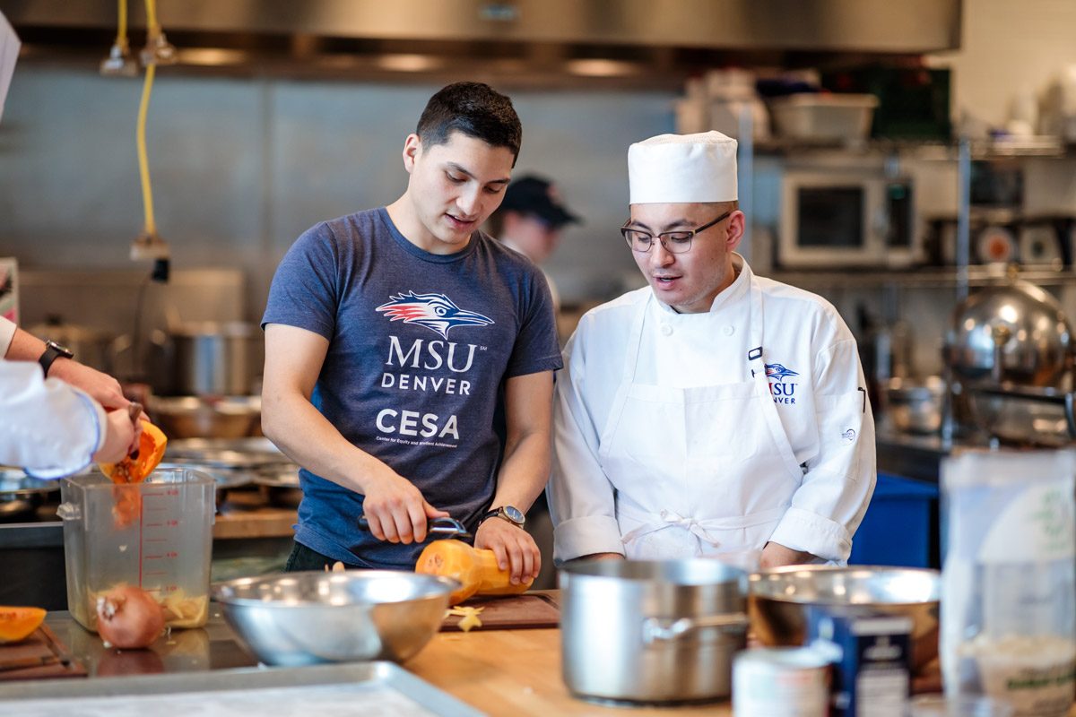 A student in a blue shirt slicing a vegetable next to a student wearing a white chef's outfit