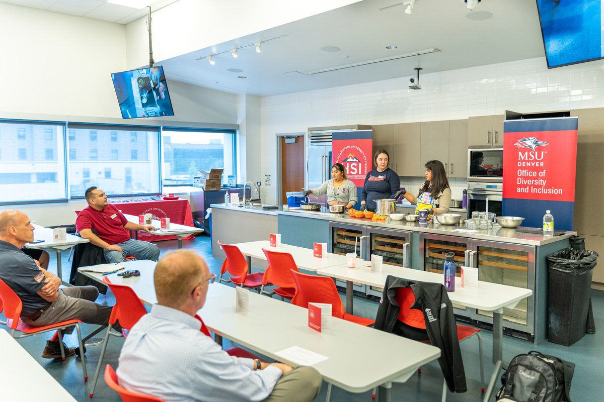 A kitchen classroom with three students presenting their food to three other people