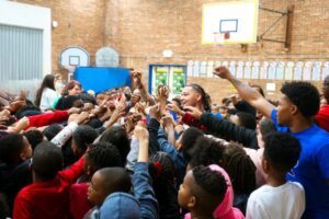 Photo of a group of MISTERs and young students standing in a school gym with their hands up and pointing to the center.