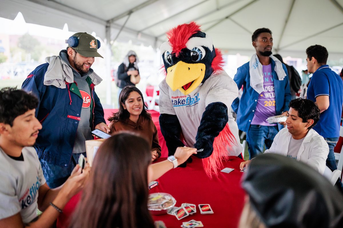 Mascot Rowdy plays Uno with a group of students gathered around a table beneath a tent outdoors