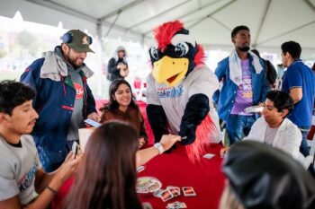 Mascot Rowdy plays Uno with students gathered around a table under a tent outdoors