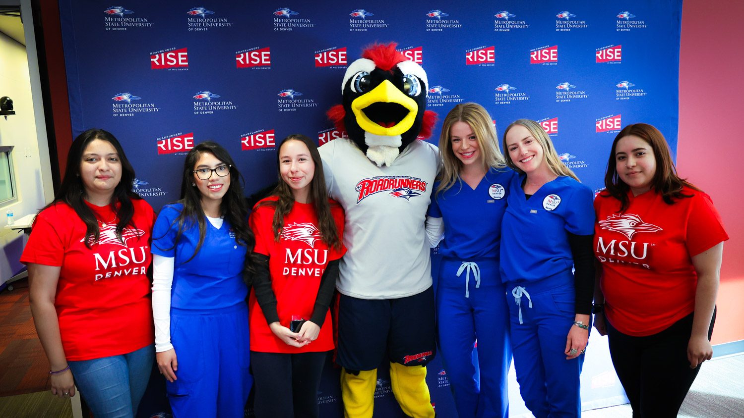 Six female healthcare students pose for a photo with Rowdy in front of a blue MSU Denver media backdrop