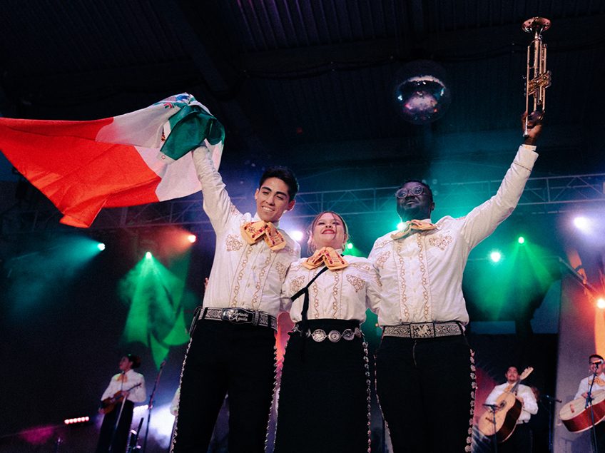 three mariachi performers waving to audience with trumpet and Mexican flag