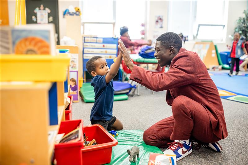 A photo of a MiSTER kneeling on the floor of a classroom, high fiving a young student.