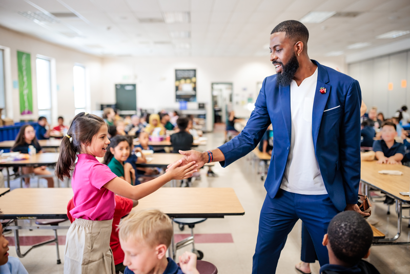Dr. Rashad Anderson exchanging a handshake with a young female student in an elementary school cafeteria.