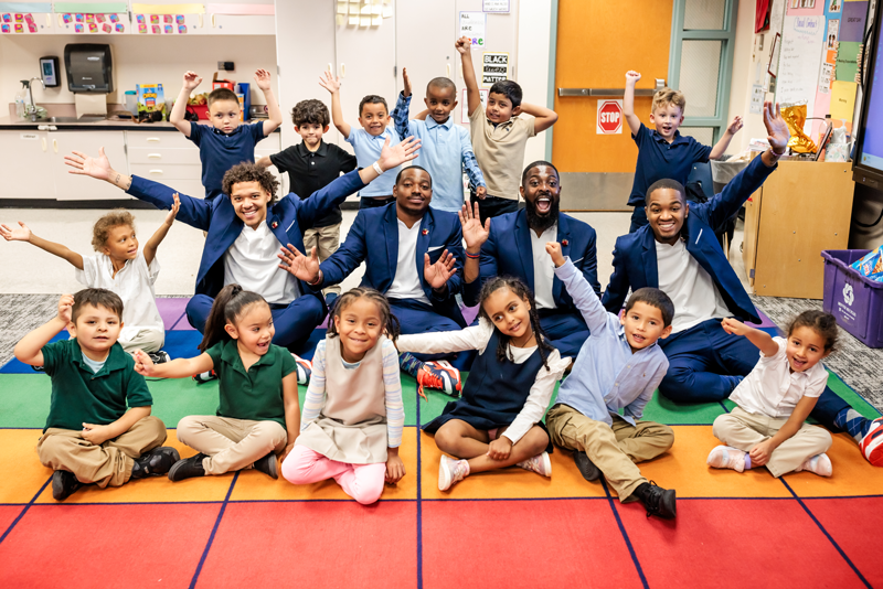 The MSU Denver Call Me MISTER students and director sit with a group of elementary school students on a colorful floor mat, arms extended and smiling.