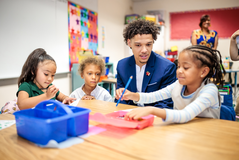 Jordan Puch, Call Me MISTER students, sits at a table in an elementary school classroom with three female students while they use colored pencils to draw.