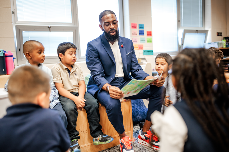 Dr. Rashad Anderson, Call Me MISTER program director, sitting on a bench in an elementary classroom reading to a small group of young students.