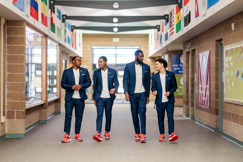 Call Me MISTER students Chris Livingston, Josh Barringer and Jordan Puch, along with Call Me MISTER program director Dr. Rashad Anderson, walking down the hallway of an elementary school toward the camera, wearing matching suits and sneakers, talking to one another.