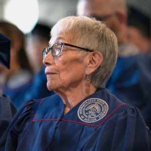 Photo of Anna Jo Garcia Haynes in MSU Denver regalia sitting watching the ceremony at the Spring 2023 Master of Arts in Teaching Hooding Ceremony.