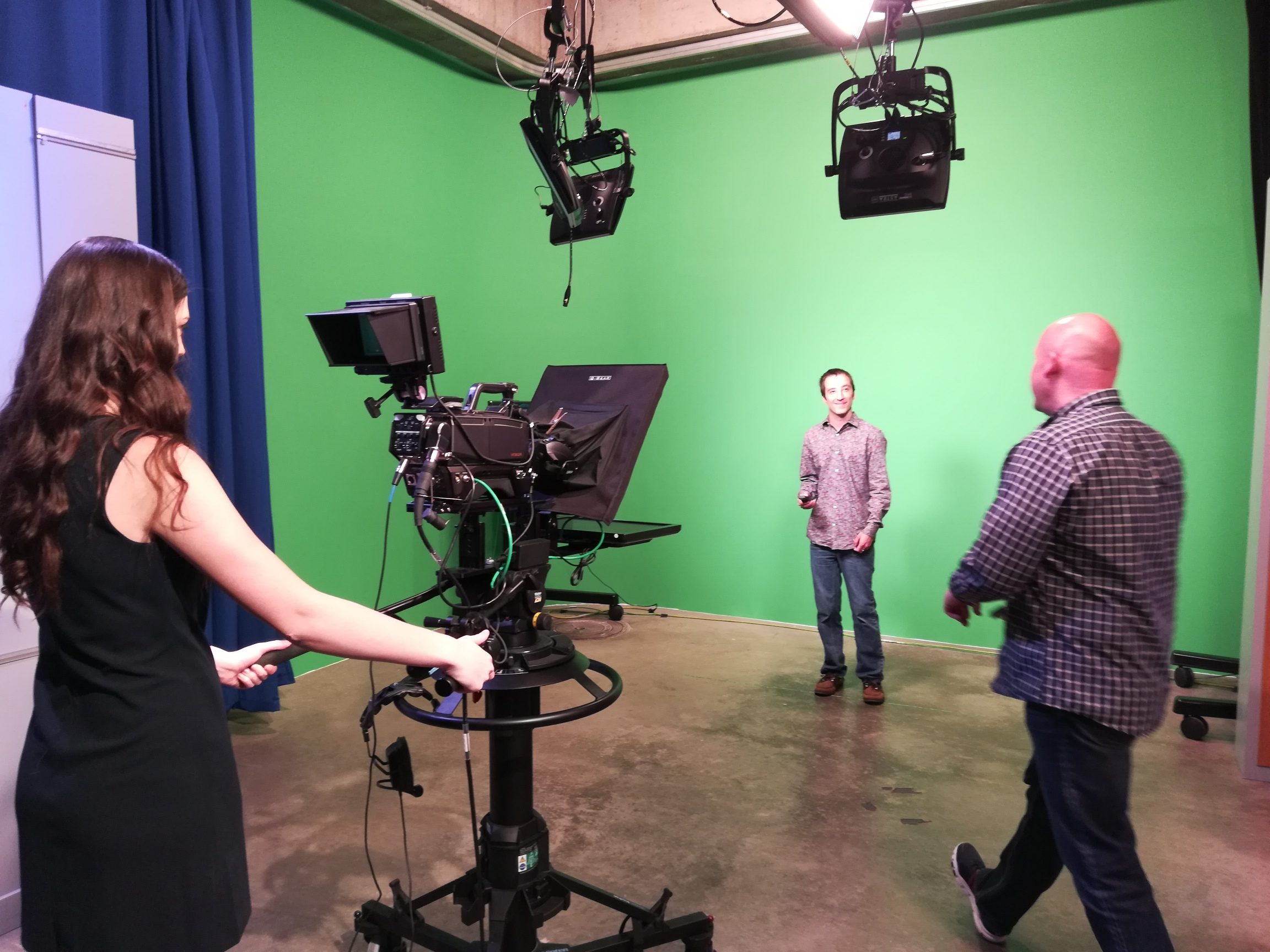 Decorative: Image of student operating a tv camera. One student stands in front of a green screen. Another student strolls by.