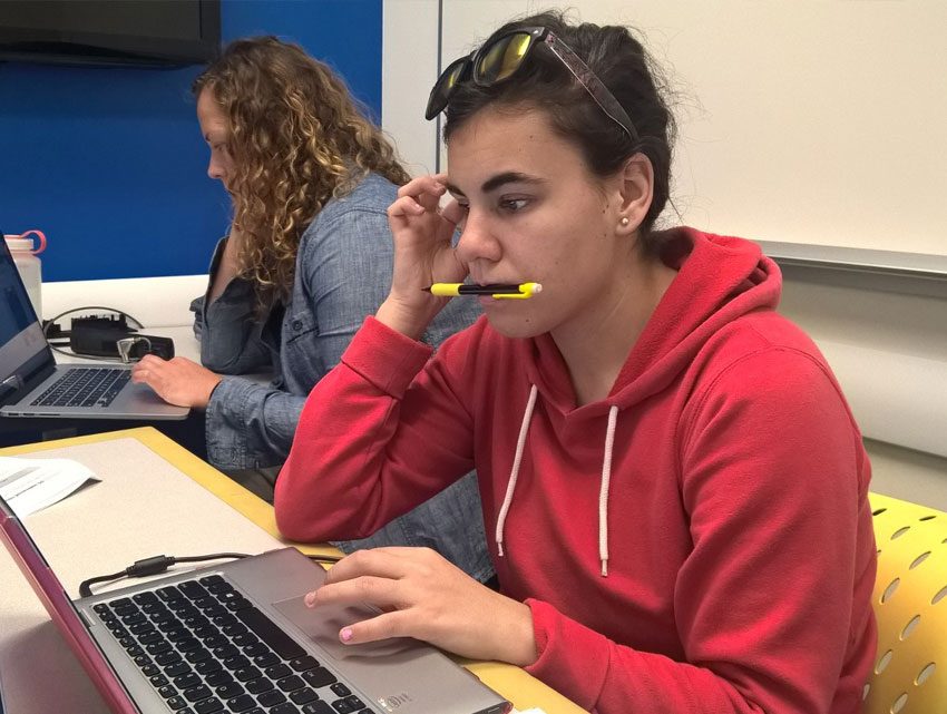Decorative: A student sits working at a laptop. They hold a pen in their mouth as they focus.