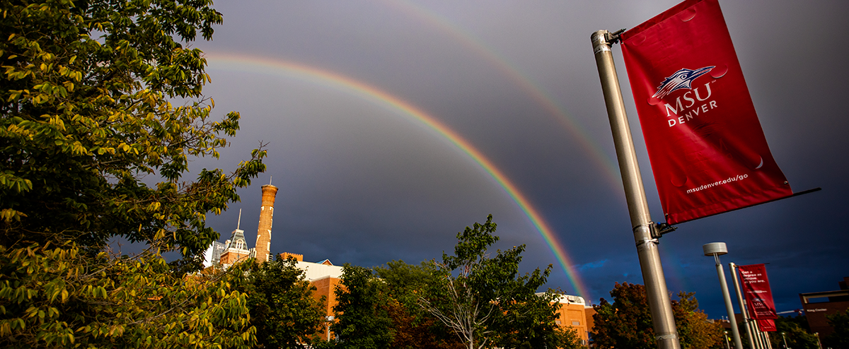 Photograph of a double rainbow above the Tivoli building with a red MSU Denver flag in the foreground.