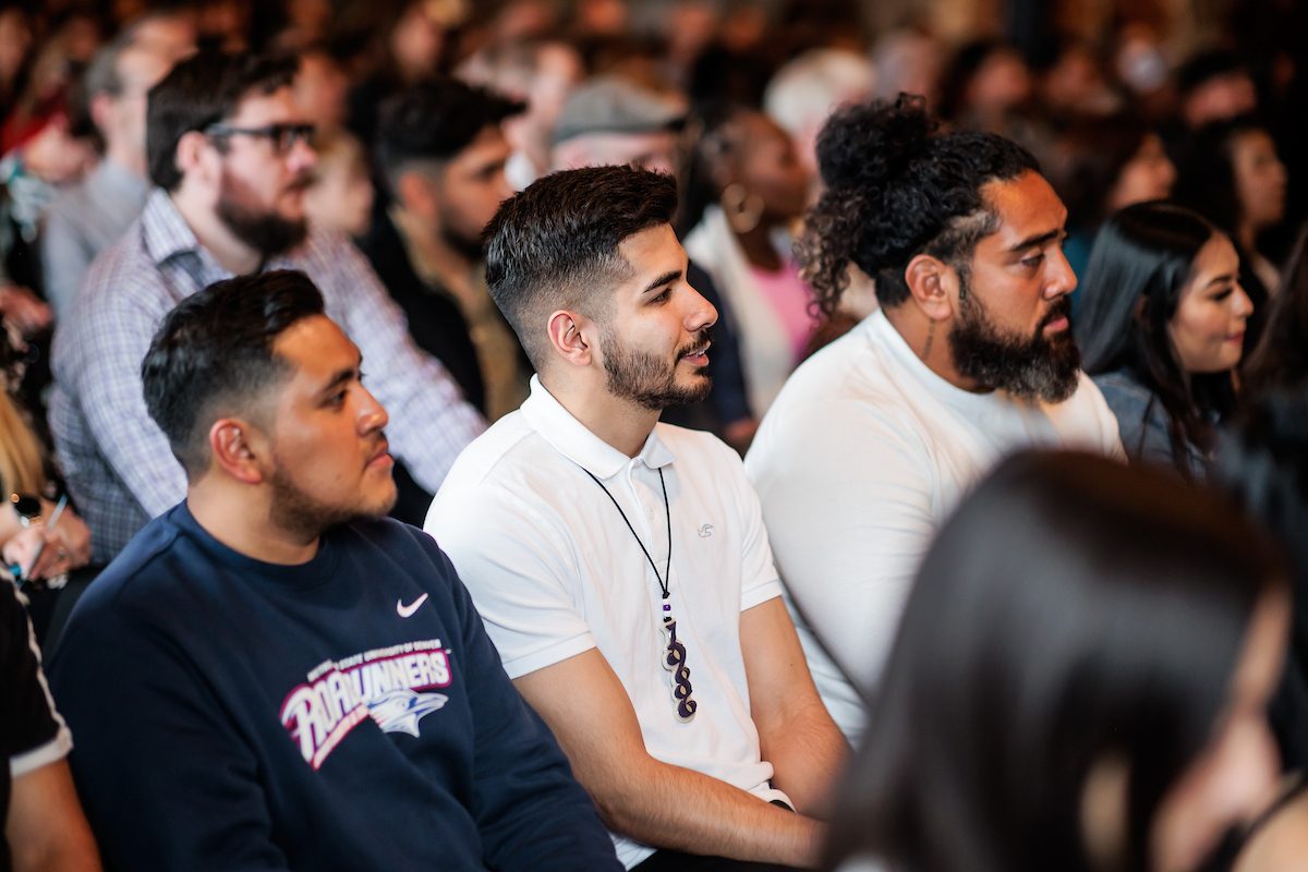 three male students of color sitting in a row of seats at an event slightly smiling
