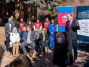 Governor Jared Polis standing at a lectern surrounded by bipartisan legislators and MSU Denver students on historic 9th Street parkway