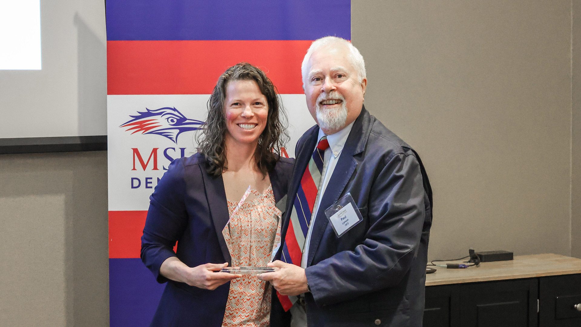 Paul Larson ('72) standing with Brandi Rideout and holding a glass award at the 2023 Golden Nest Club luncheon