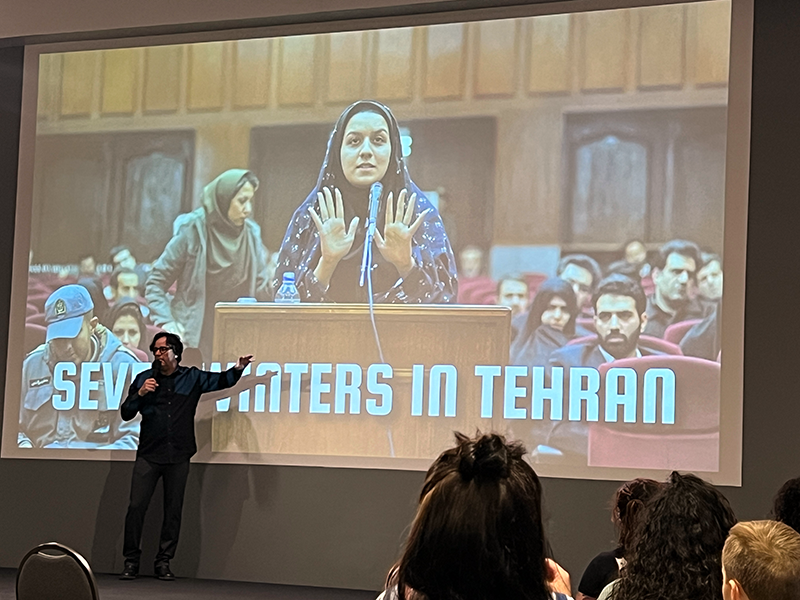 The image shows Dr. Vincent Piturro speaking in front of a projection screen displaying a still from the film 