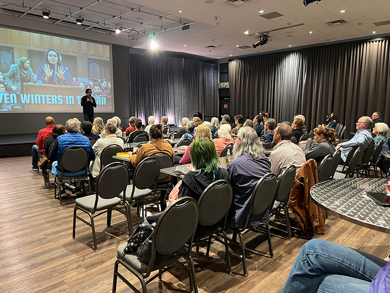 The image shows Dr. Vincent Piturro, professor of film and media studies at MSU Denver, speaking into a microphone while standing in front of a large audience seated in rows of chairs. Behind Dr. Piturro, a projection screen displays the title 