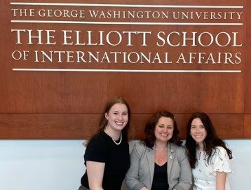 Three students sitting under a sign that says: The George Washington University, The Elliot School of International Affairs
