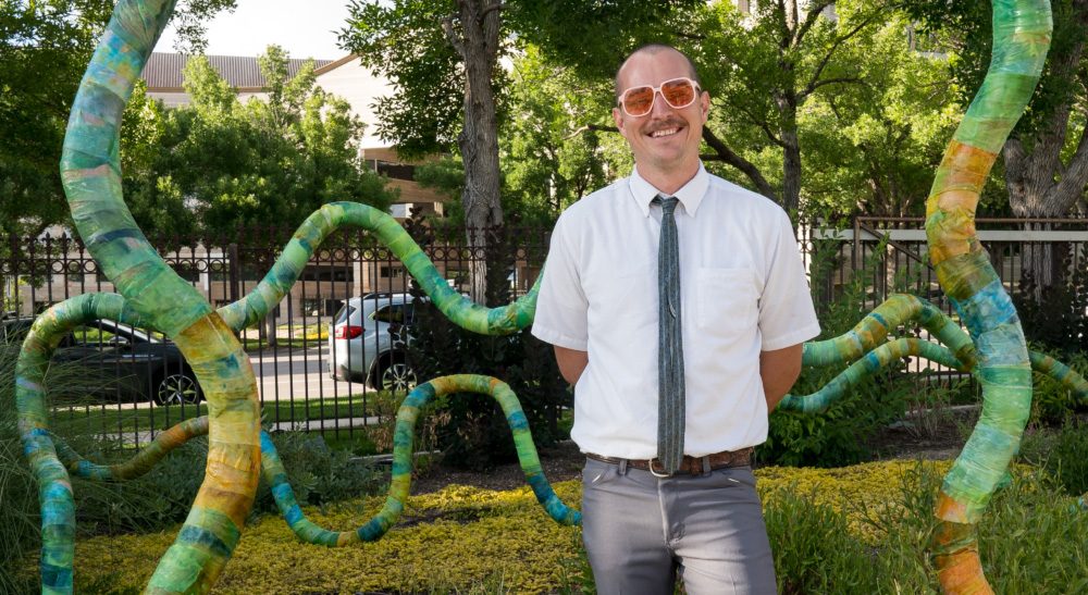 Charles, shown here in front of a giant outdoor sculpture, wiggle-like shape of mulicolored greens, blues, and yellows, has been named summer 2024 MOA Design and Build Artist Fellow.