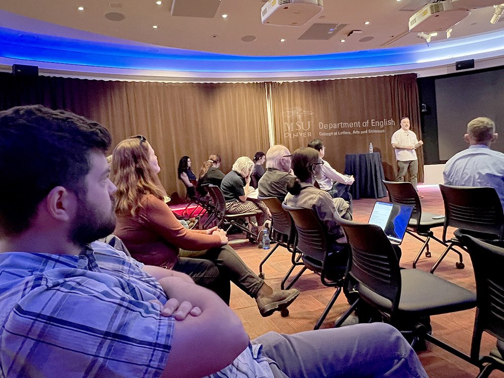 The image shows the audience perspective from a presentation by Travis Masingale from Eastern Washington University about using AI in college classrooms. The presentation is taking place in the MSU Denver CAVEA and sponsored by the Department of English at Metropolitan State University Denver. A room full of attendees, primarily faculty members, are seated in curved rows facing the presenter, with laptops and notebooks in front of them for taking notes. Professor Masingale is standing at the front of the room delivering his talk.