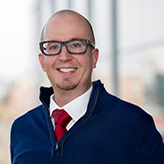 Headshot of Cory Phare, wearing a blue sweater over a white button-up with a red tie and stylish (for 2016) glasses.