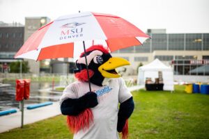 Rowdy, the MSU Denver University mascot, dressed as a roadrunner, stands on a large rock with arms raised and thumbs up. The scene is set on a grassy campus area with the Tivoli building and the Denver city skyline in the background.
