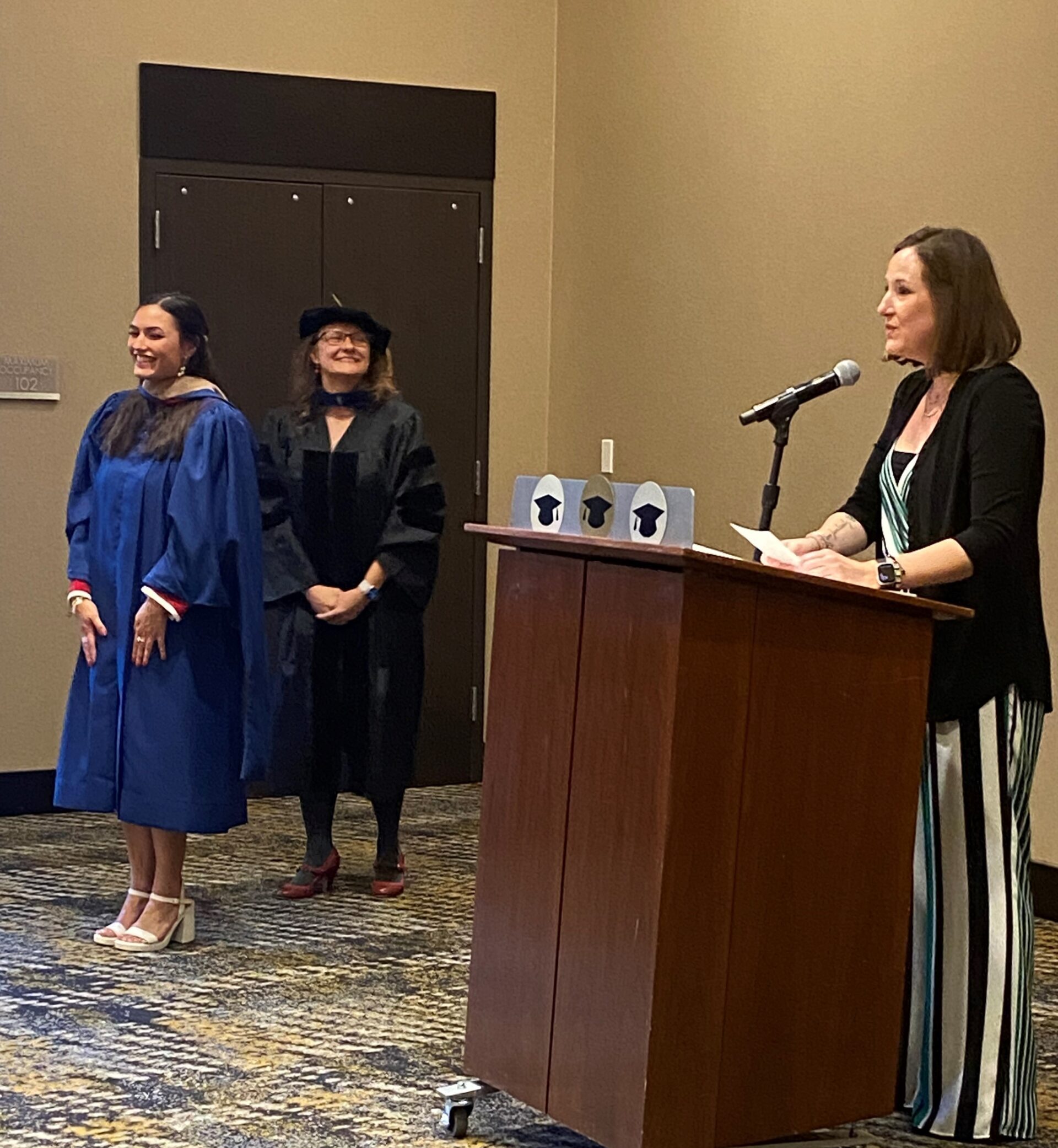 SP'24 MBA & MPAcc Hooding Celebration - Faculty (Jo) at the podium talking about graduating student, Emily, as she stands next to the podium after being hooded by faculty (Letitia)