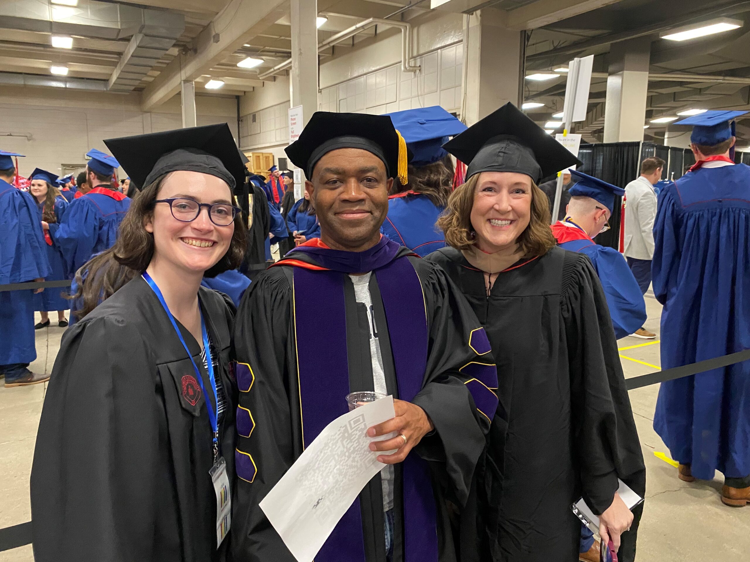 Spring 2024 Commencement - Staff (Lauren), Department Chair (Greg) & faculty (Jo) smiling in graduation regalia as they are waiting to line up and enter the coliseum main floor