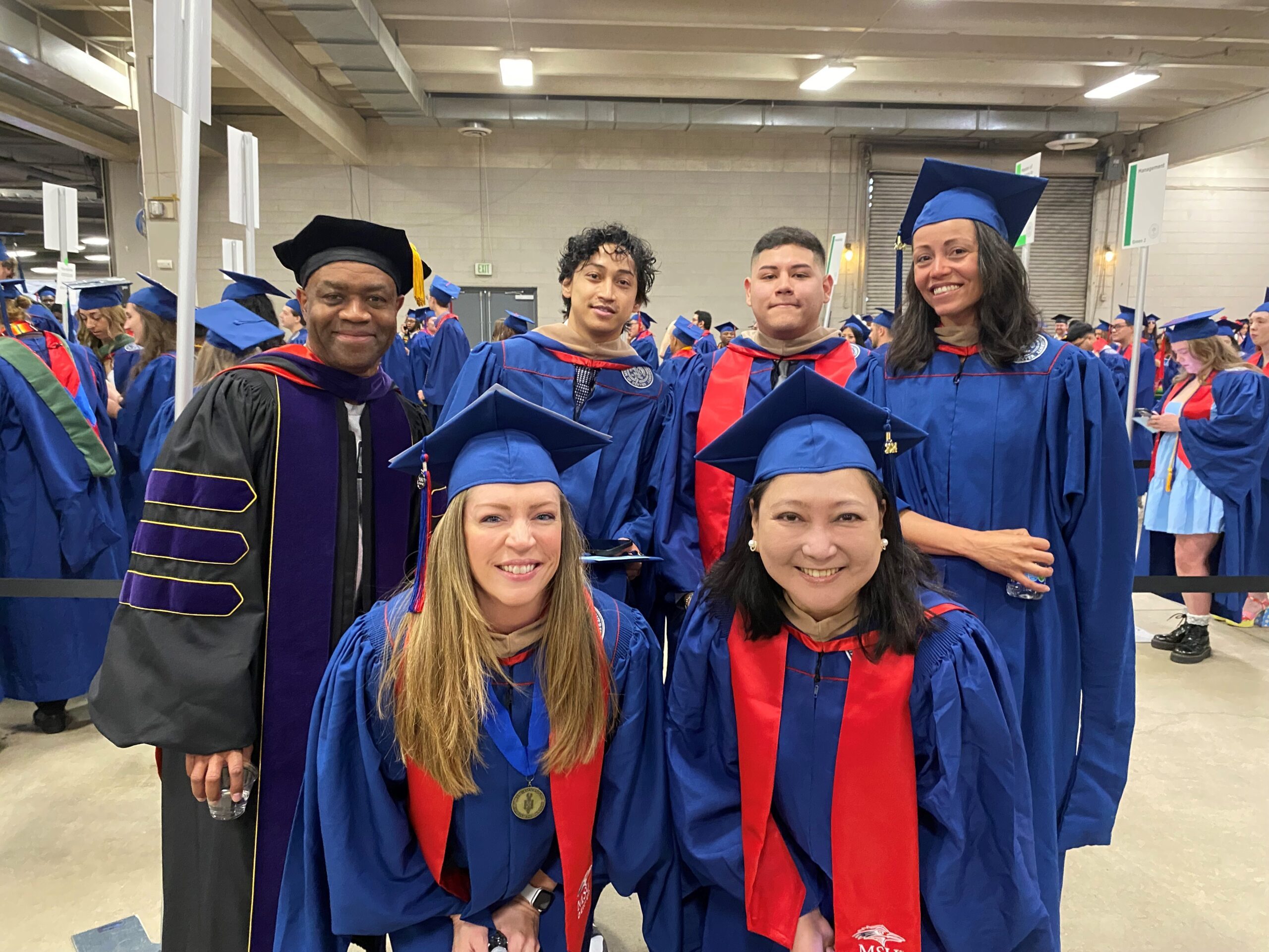 Spring 2024 Commencement - Department Chair (Greg) & 5 MPAcc students smiling in graduation regalia as they are waiting to line up and enter the coliseum main floor