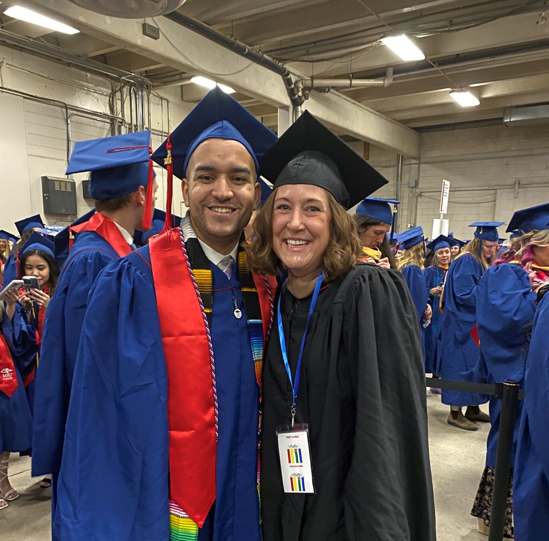 Spring 2024 Commencement - faculty (Jo) & undergraduate accounting student (Kevin Roman) smiling in regalia as they wait to line up for the commencement ceremony