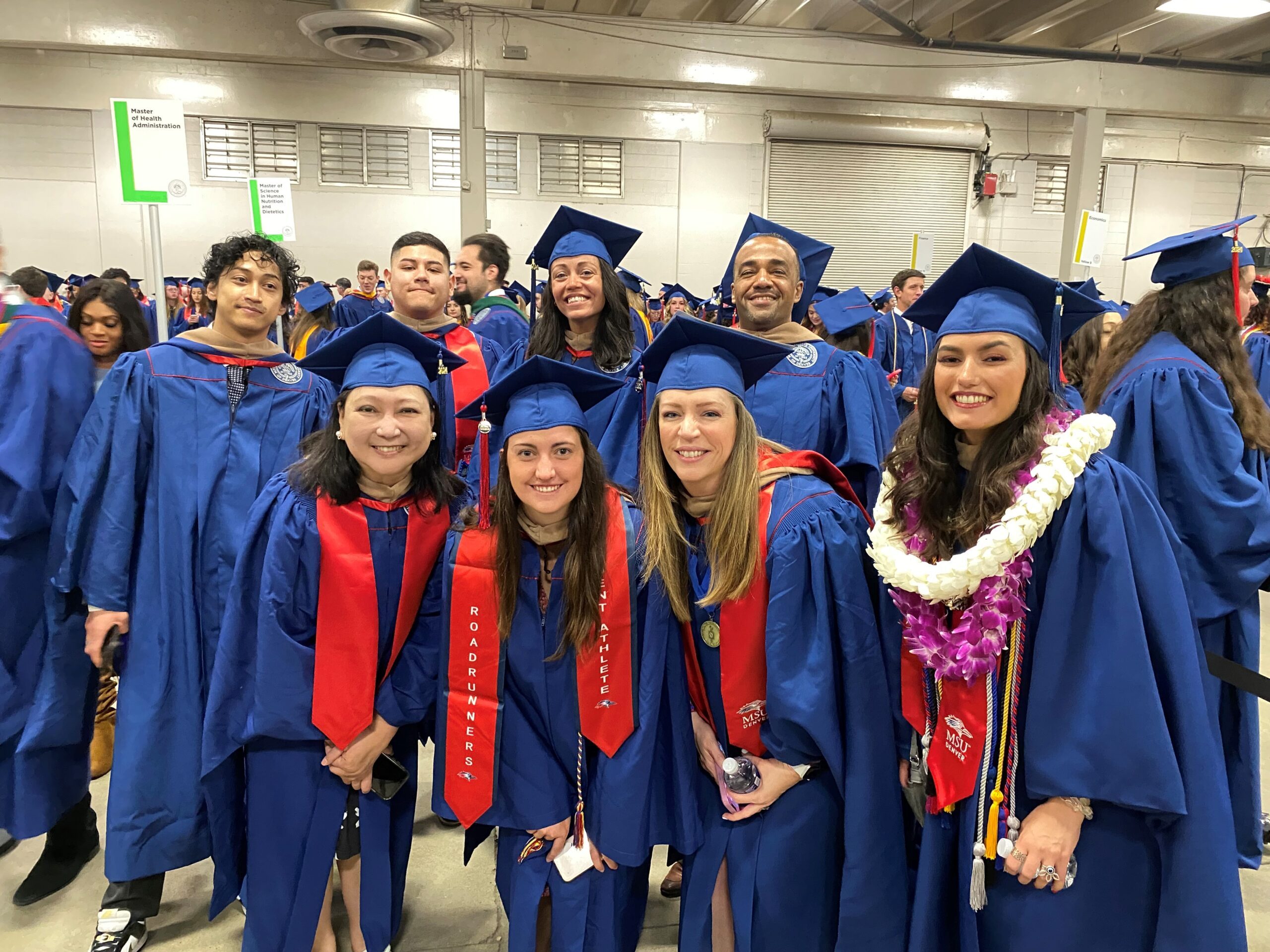 Spring 2024 Commencement - 8 MPAcc students smiling in graduation regalia as they are waiting to line up and enter the coliseum main floor