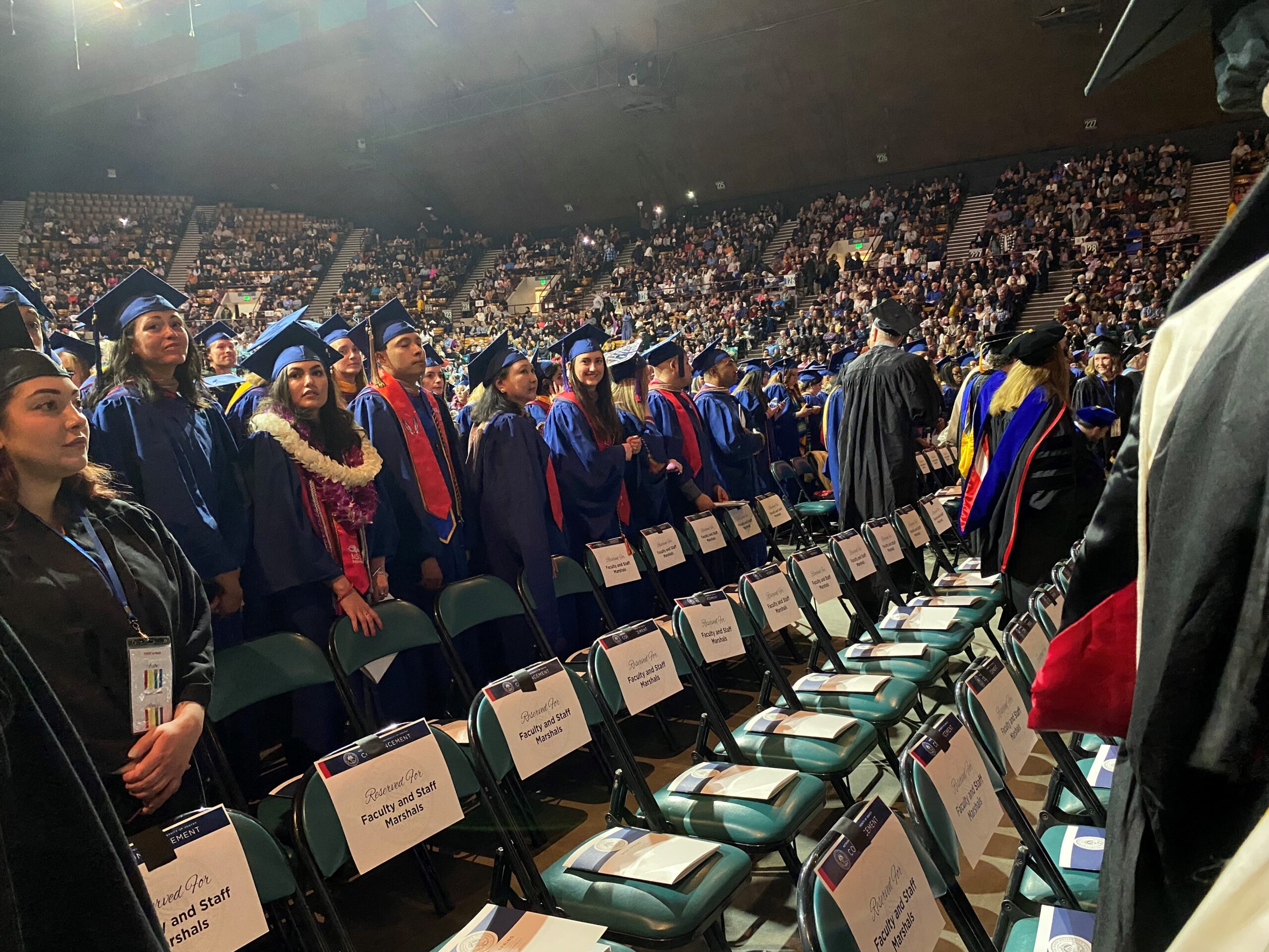 students in regalia at graduation looking at family and friends in stands as they are standing waiting to take their seats for the start of the commencement ceremony (SP'24)
