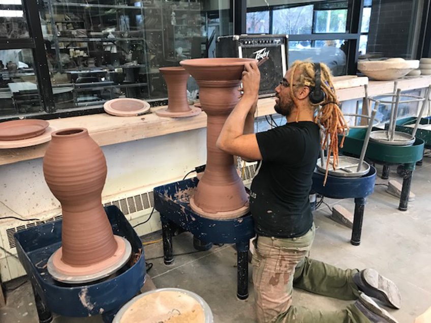 Student kneeling on floor of ceramics studio working on a large (2 feet 6 inches wheel thrown form). There is a second large wheel thrown form on the adjacent potters wheel.