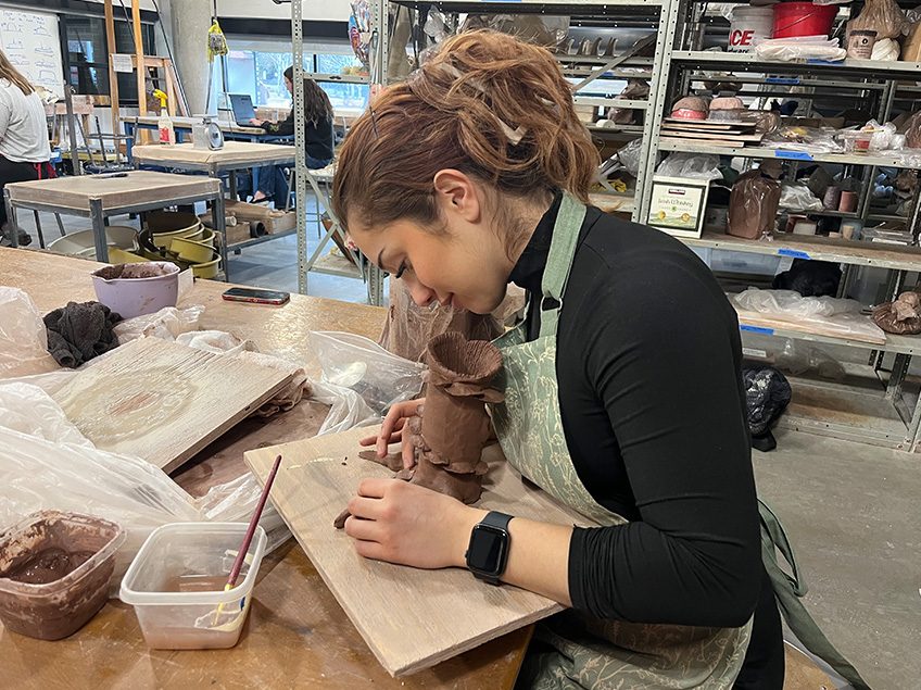 Female student working on an unfired hand built vessel in the ceramics classroom with view of storage shelves in the background .