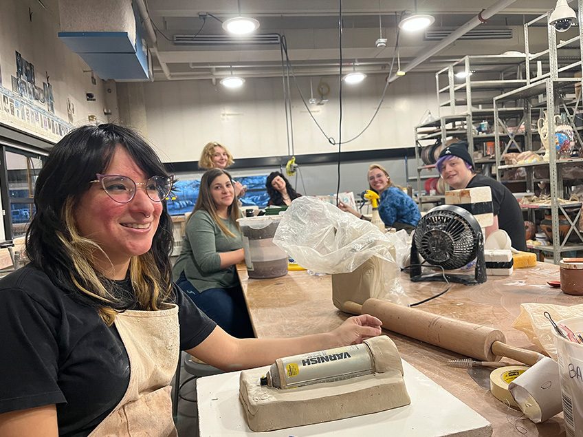 Seven students smiling for the camera as they are also making molds and slip casting in the ceramics classroom with view of the storage shelving and windows in the background.
