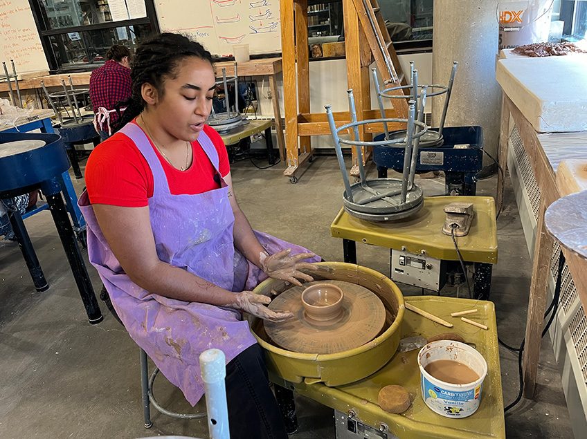 Student sitting at a potters wheel throwing a small bowl with a view of additional potter’s wheels in the background in the ceramics classroom.