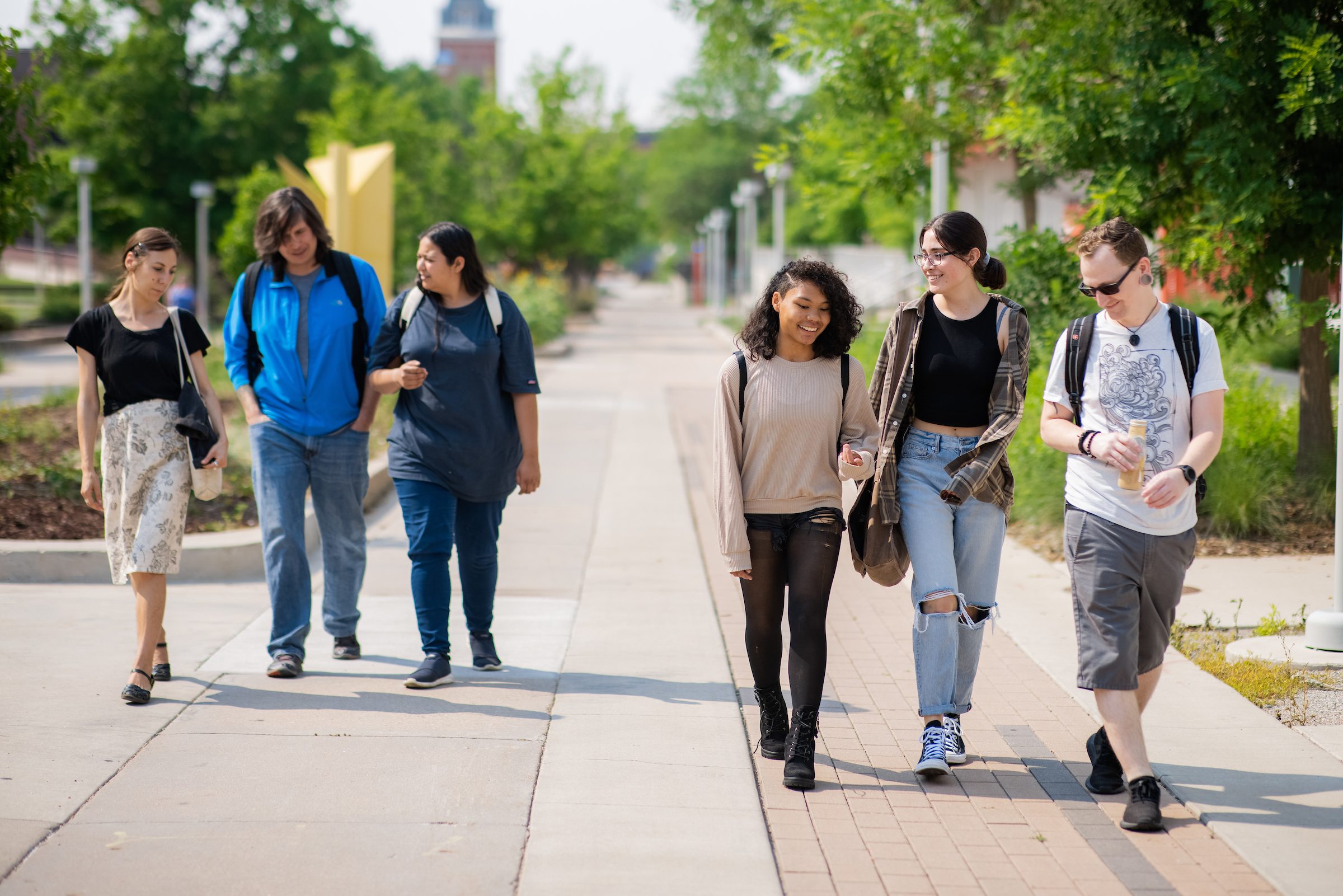 A group of six MSU Denver students walking together on campus on a sunny day. The students are dressed in casual attire, chatting and smiling as they walk. The background shows a tree-lined pathway with the campus buildings in the distance.