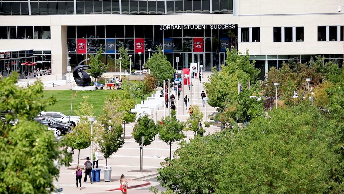 An aerial view of the MSU Denver campus, focusing on the Jordan Student Success Building. The entrance is busy with students walking along the pathways lined with trees. The building has large glass windows and 'Jordan Student Success' is written above the entrance. A sculpture is visible on the lawn, and a coffee shop with outdoor seating is on the left.