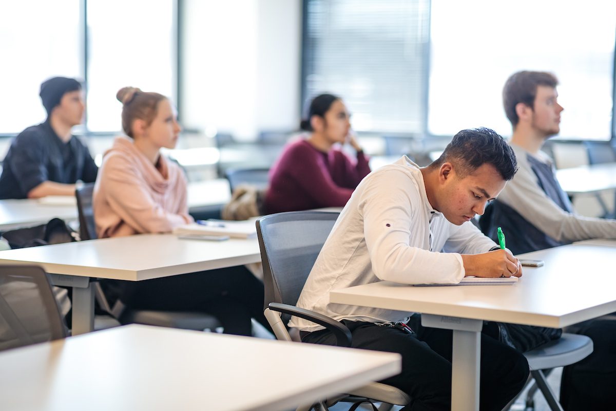 Several students sitting at desks in a classroom. The focus is on a student in the foreground wearing a white shirt and writing with a green pen. Other students in the background are attentively listening and taking notes.