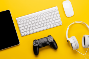 Image of laptop, headphones, game controller, mouse and iPad on yellow background.
