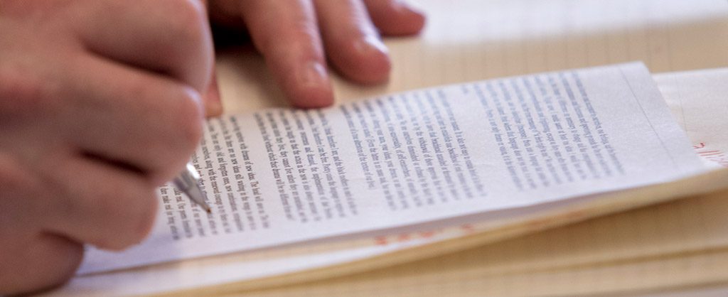 A close-up of a person's hand writing on a small piece of paper with printed text, resting on a larger lined notepad.