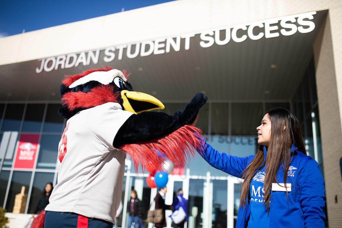 MSU Denver's mascot, Rowdy the Roadrunner, giving a high-five to a student in front of the Jordan Student Success Building. Rowdy is dressed in a white jersey, while the student is wearing a blue MSU Denver jacket. The background shows the building's entrance and signage.
