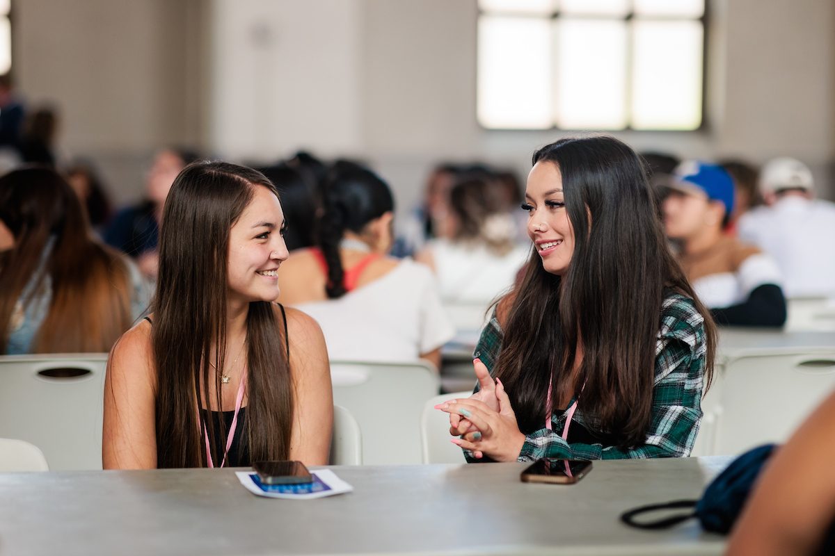 Two female students sitting at a table, smiling and talking to each other during an orientation event. The background shows other students seated and engaging in conversation.